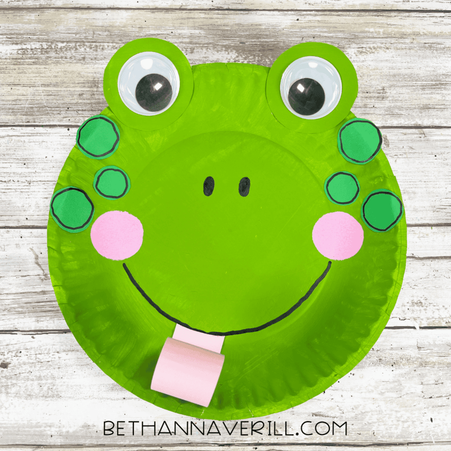 A finished frog paper plate craft with big googly eyes, green spots, rosy cheeks, a smile, and a curly pink tongue.