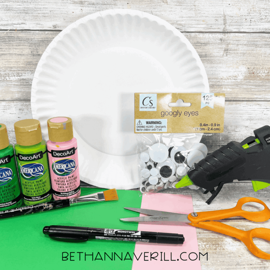 Craft supplies including a paper plate, green and pink acrylic paint, googly eyes, scissors, glue gun, and construction paper laid out on a table.