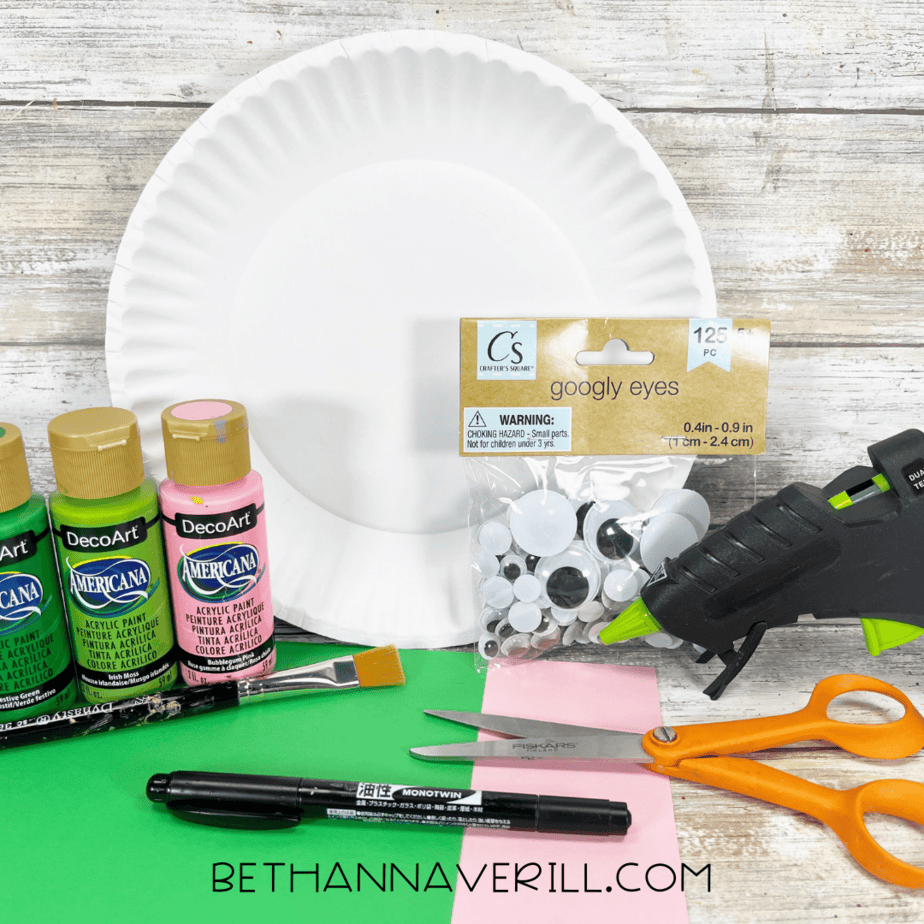Craft supplies including a paper plate, green and pink acrylic paint, googly eyes, scissors, glue gun, and construction paper laid out on a table.