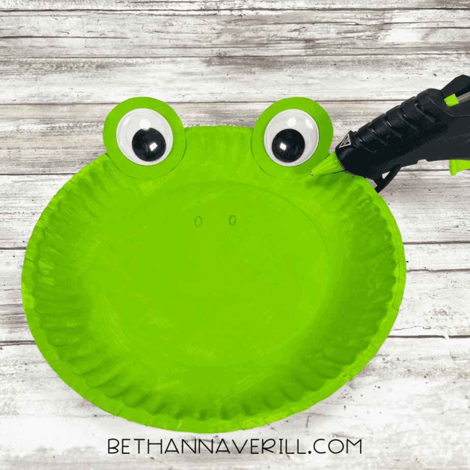 A green paper plate with two large googly eyes glued on top of green circles at the rim.