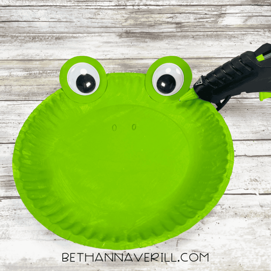 A green paper plate with two large googly eyes glued on top of green circles at the rim.