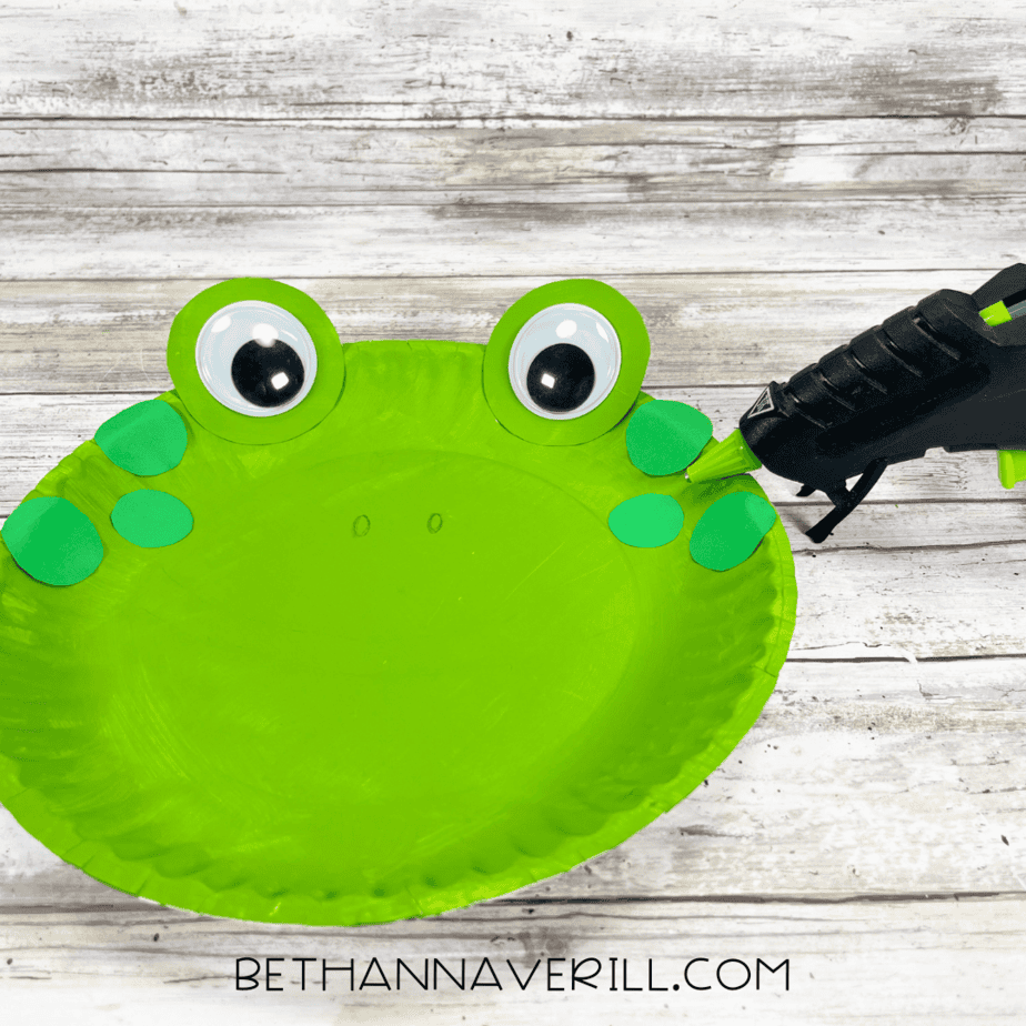 A glue gun attaching small green circles along the sides of the frog’s face for spots.