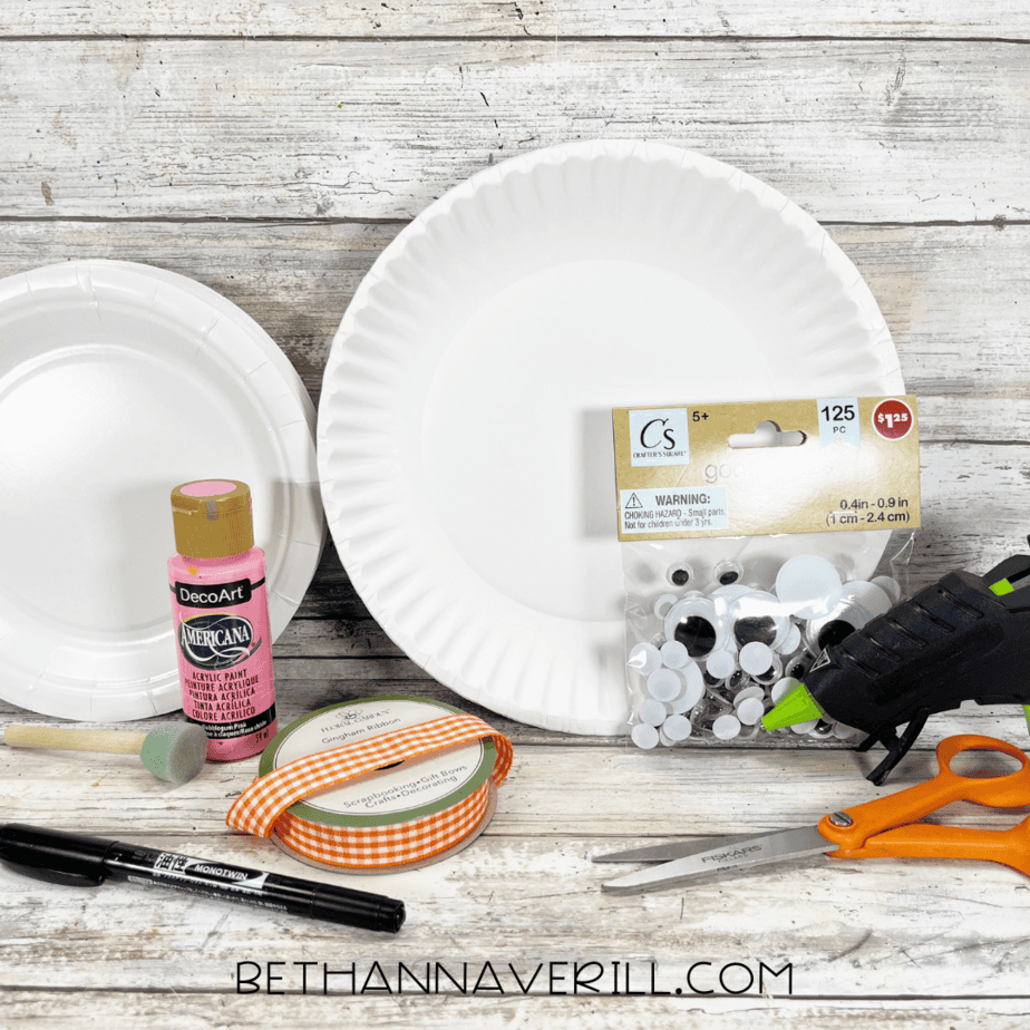 Craft materials laid out including paper plates, pink paint, ribbon, googly eyes, scissors, marker, glue gun, and paint sponge.