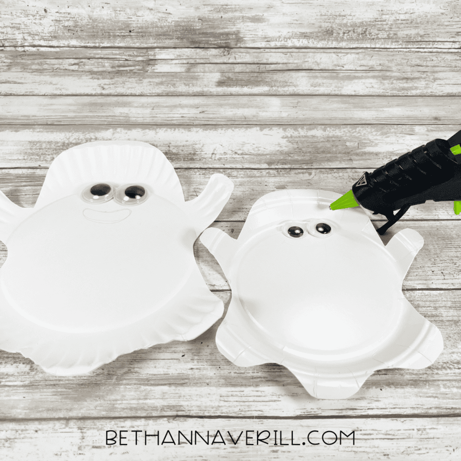 Large and small paper plate ghosts with googly eyes glued on, one with a glue gun beside it.