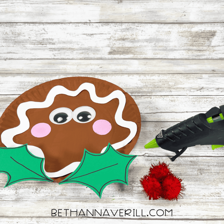 Holly leaves placed under the gingerbread face with glue gun and red pom poms nearby.