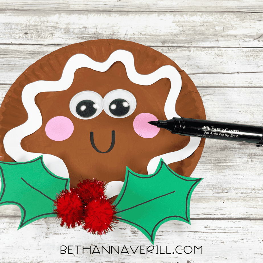 Gingerbread paper plate with holly leaves and red pom poms glued in place, marker adding a smile.
