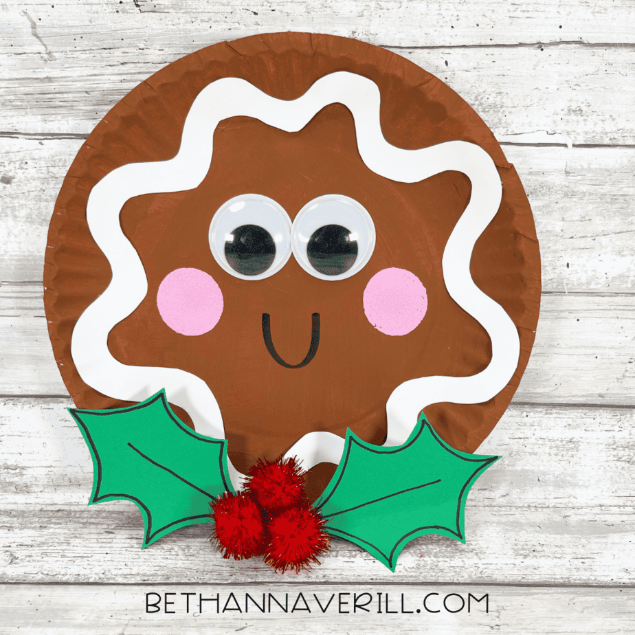 Completed gingerbread paper plate craft with googly eyes, pink cheeks, icing trim, and holly decoration.