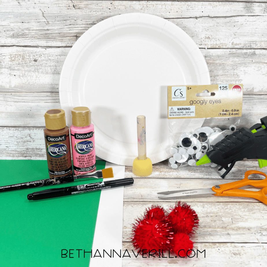 Craft supplies including paper plate, brown and pink paint, paintbrushes, googly eyes, glue gun, scissors, and red pom poms.