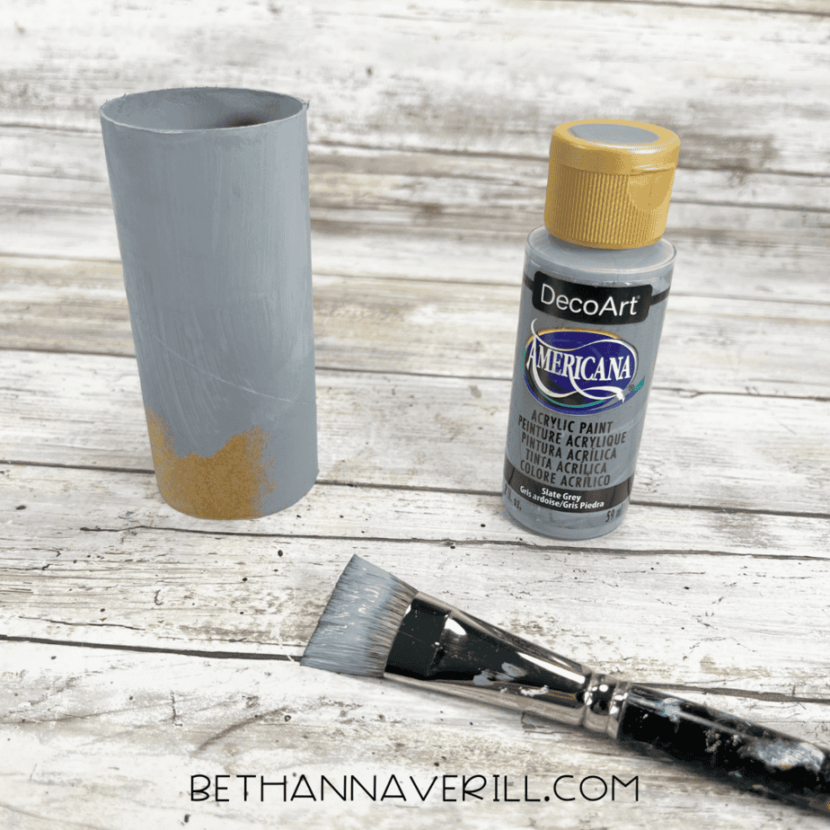 Toilet paper roll painted gray with gray paint bottle and paintbrush nearby.