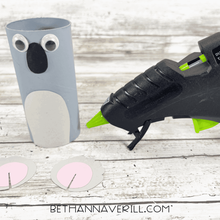 Toilet paper roll with googly eyes, black nose, and gray belly glued on, glue gun beside.