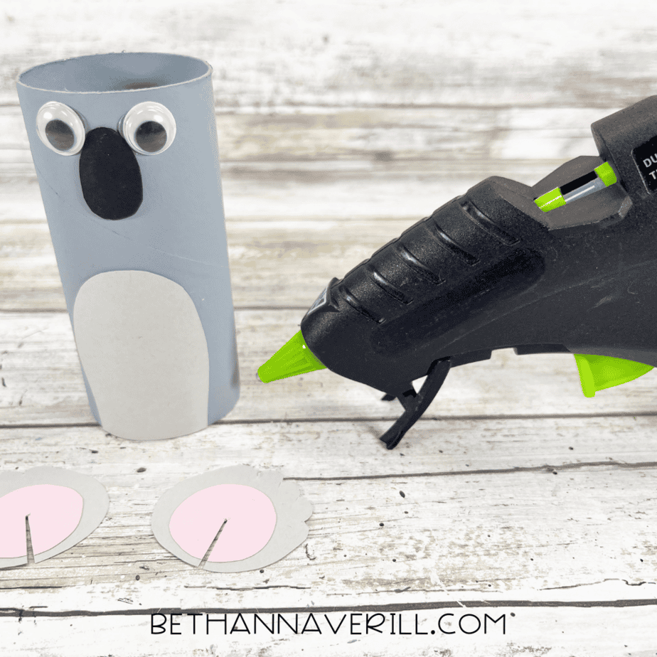 Toilet paper roll with googly eyes, black nose, and gray belly glued on, glue gun beside.
