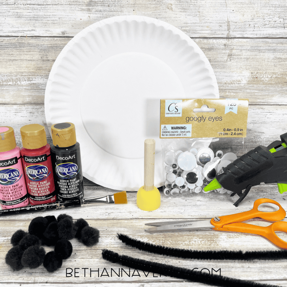 Craft supplies for a paper plate ladybug including paints, pom-poms, pipe cleaners, googly eyes, and a paper plate.