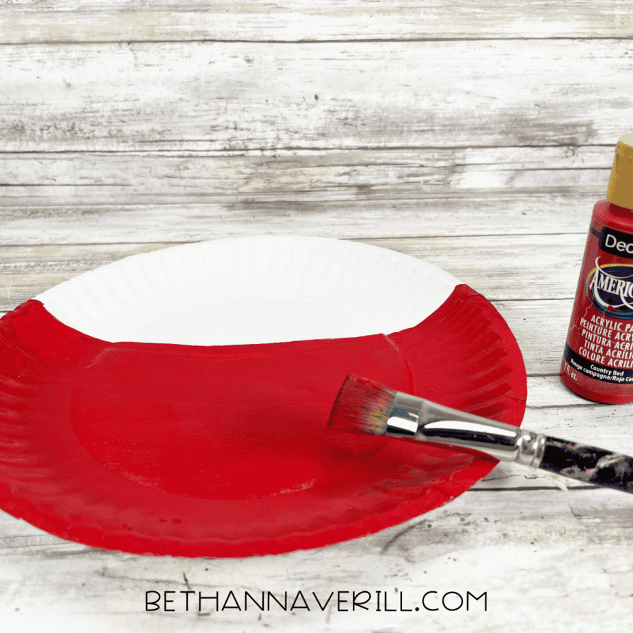 Paper plate being painted red with a paintbrush.
