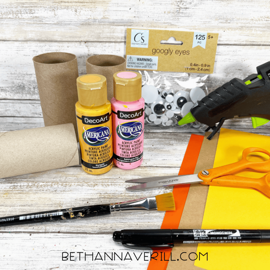 Materials for lion paper roll craft including paint, paper, googly eyes, and glue gun.