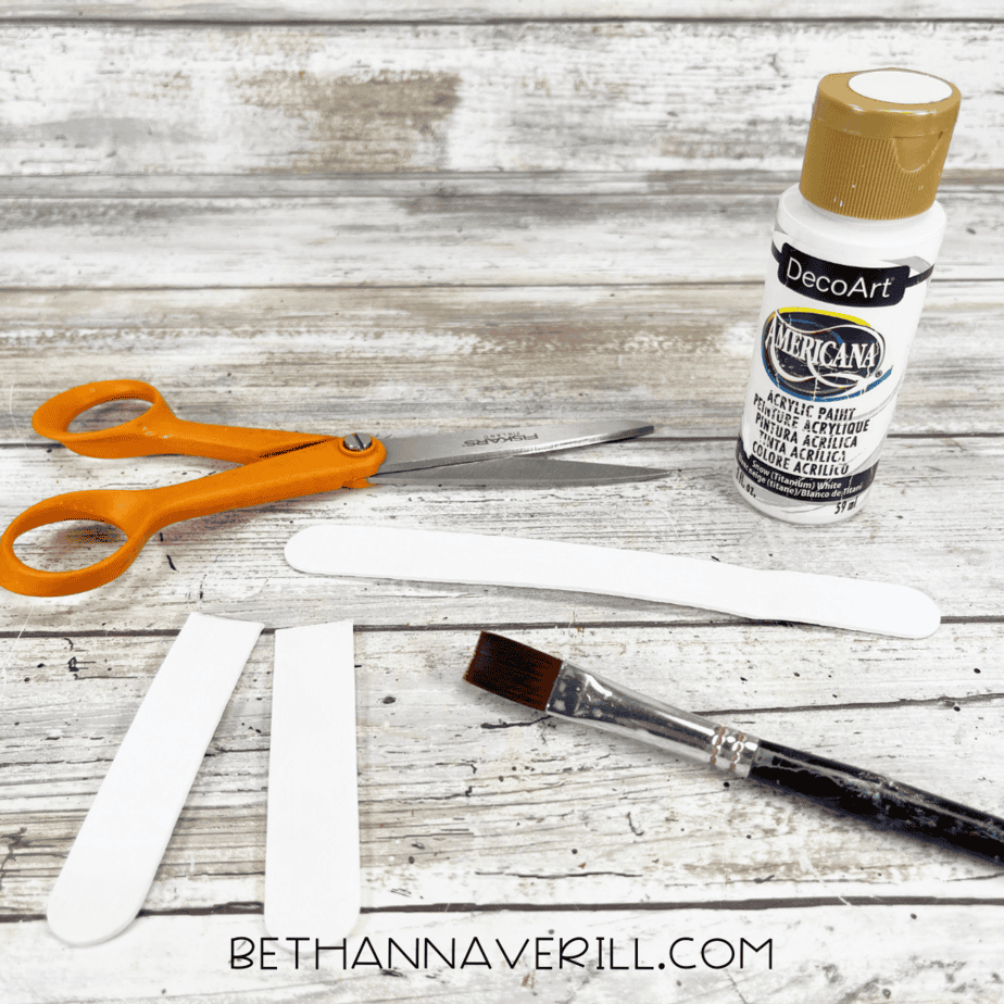 Four craft sticks painted white and cut for the llama’s legs, shown with scissors, paint, and a brush.