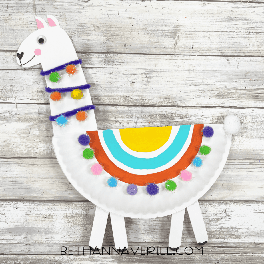 A finished paper plate llama decorated with a rainbow saddle, pom-poms, googly eyes, pipe cleaner, and painted legs.