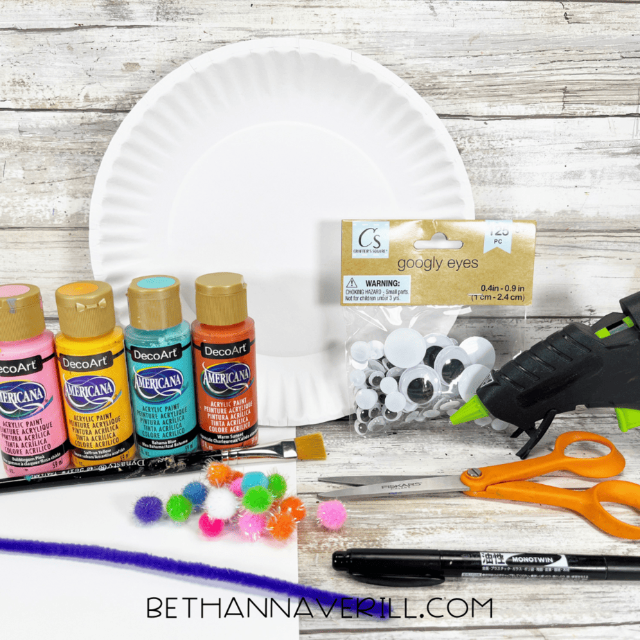 Supplies for llama paper plate craft including paints, pom-poms, googly eyes, and paper plate.