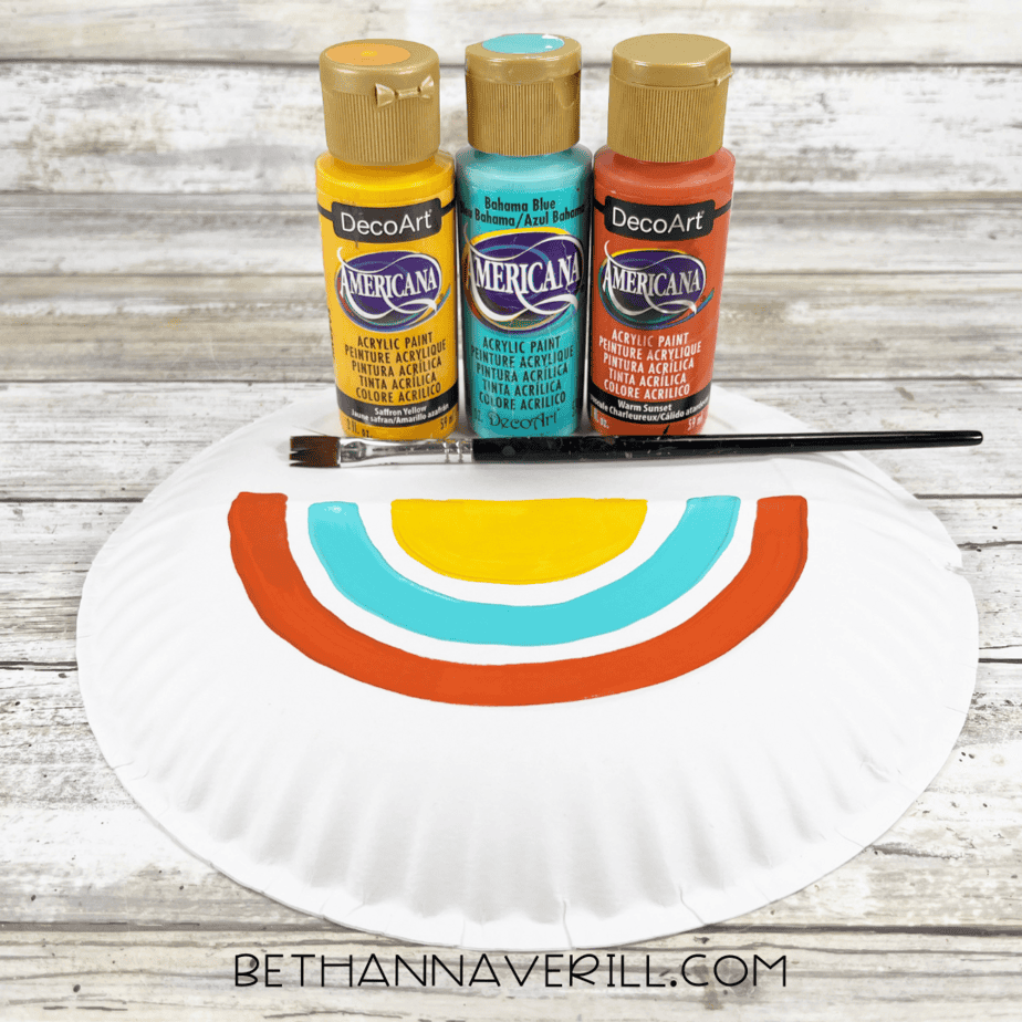 Half of a paper plate painted with rainbow-colored arches in yellow, turquoise, and orange with paint bottles nearby.