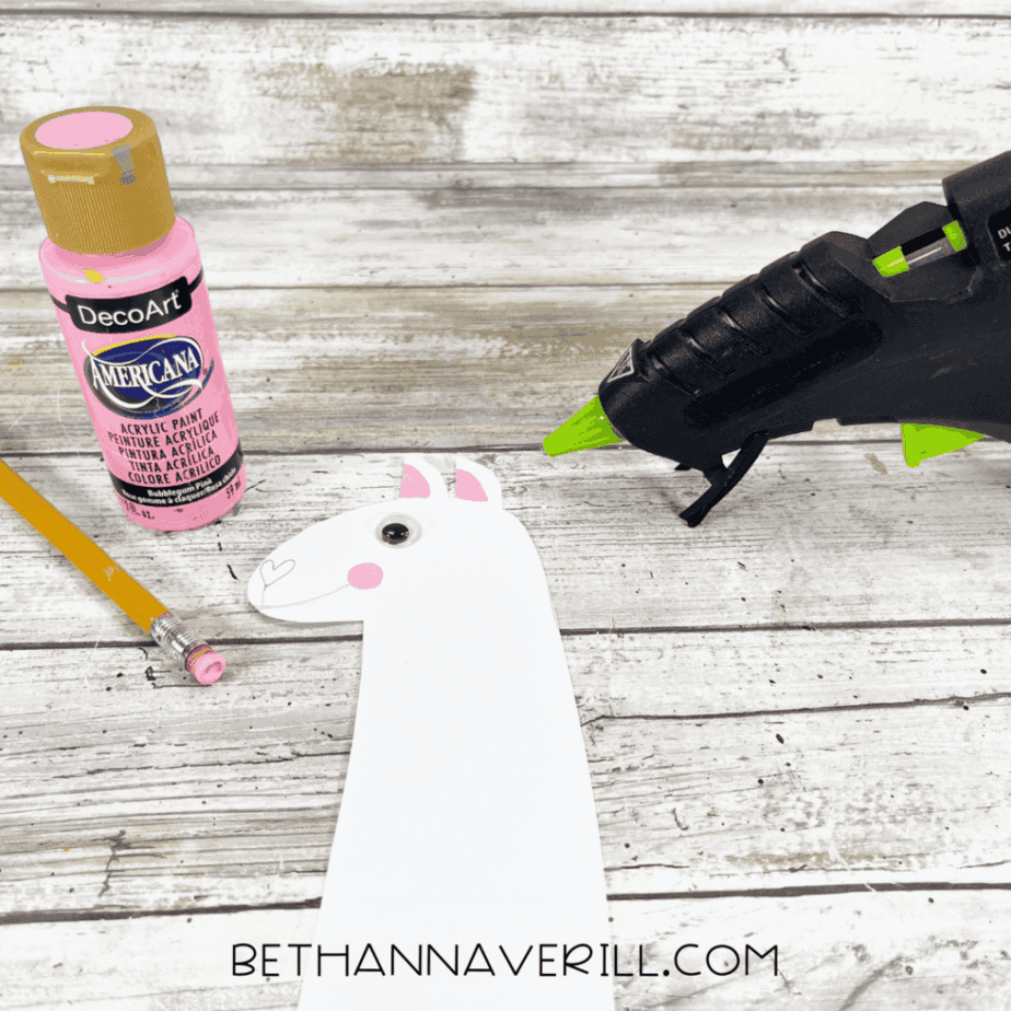 The cardstock llama head decorated with a googly eye, pink cheeks, and inner ears, shown next to a glue gun and pink paint.