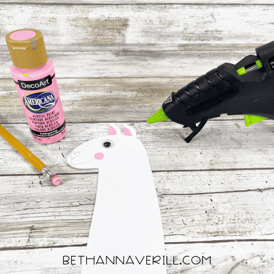 The cardstock llama head decorated with a googly eye, pink cheeks, and inner ears, shown next to a glue gun and pink paint.