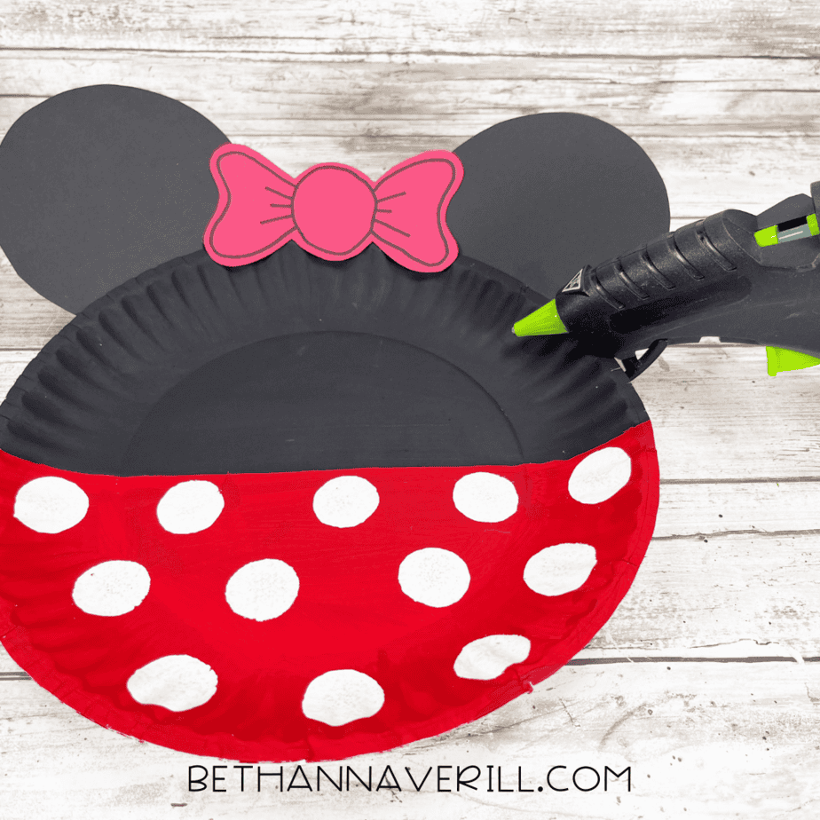 Paper plate with red bow being glued on between Minnie’s ears.