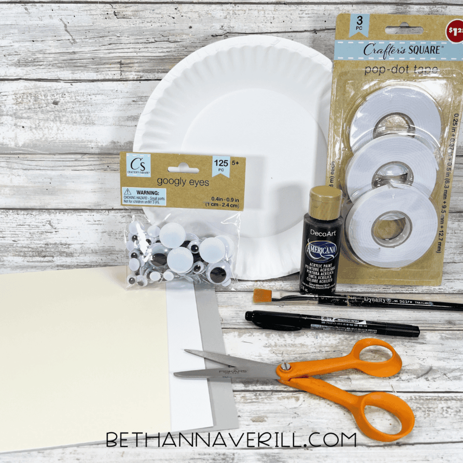 Craft supplies for mummy paper plate craft including paper plate, googly eyes, black paint, construction paper, scissors, and glue.
