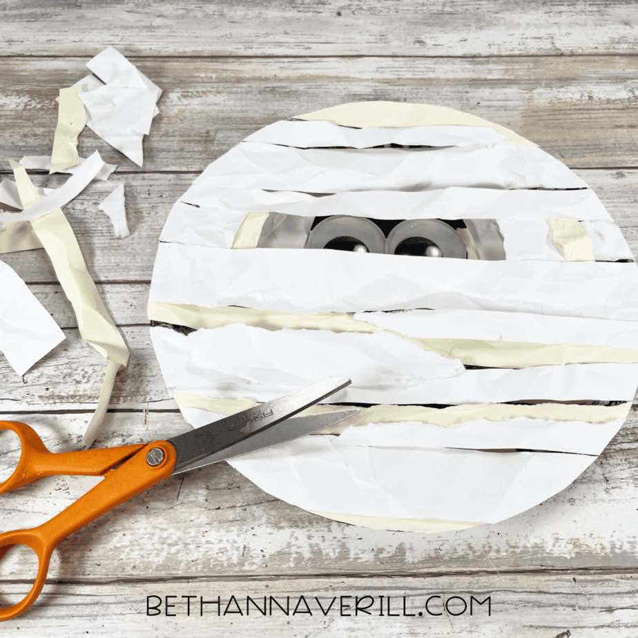 Paper plate mummy with excess strips being trimmed using scissors.