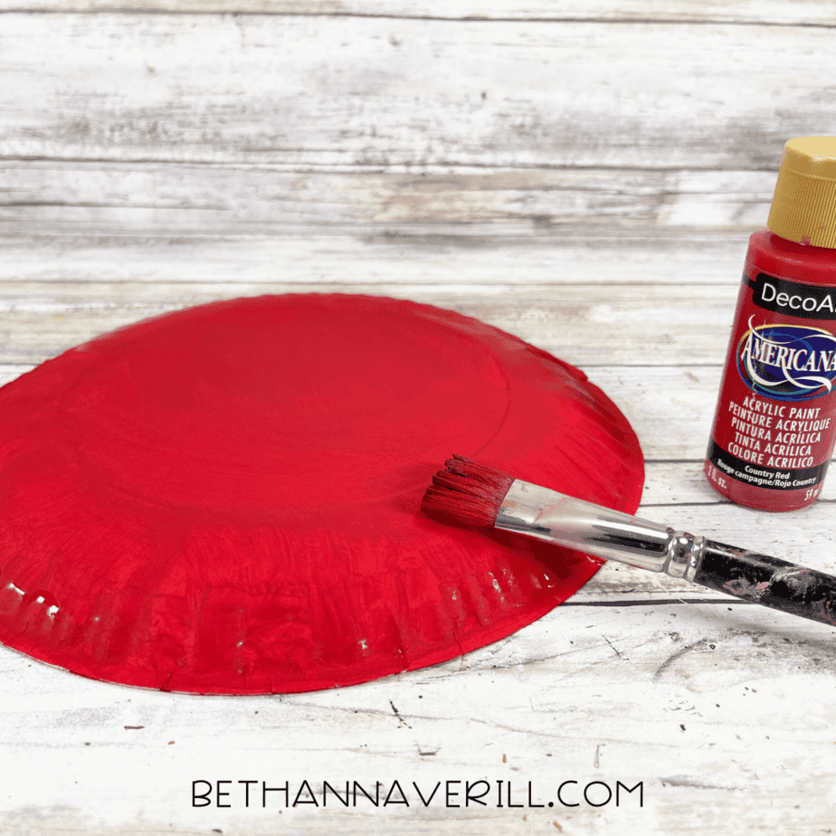 Red paint being brushed onto the back of a paper plate.