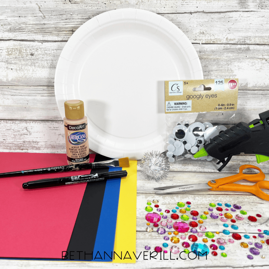 Materials for Nutcracker paper plate craft including paint, paper plate, googly eyes, construction paper, gems, scissors, and glue gun.