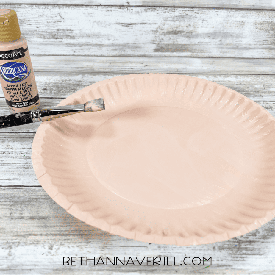 Paper plate painted with peach skin-tone acrylic paint and paintbrush beside it.