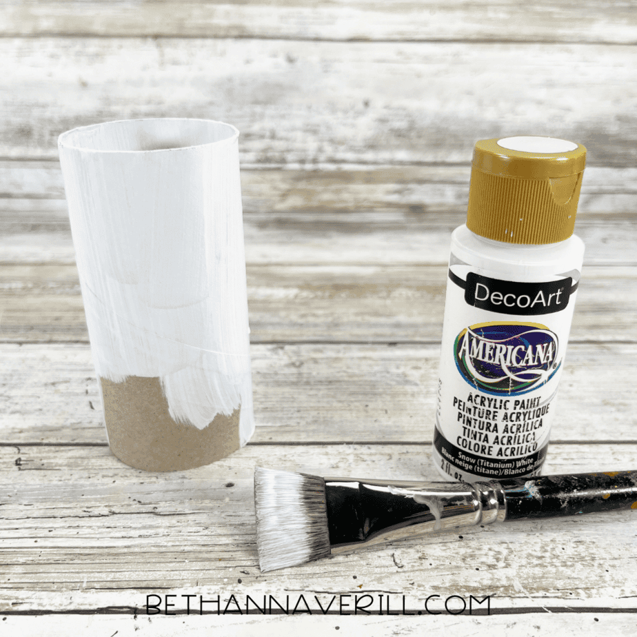 Toilet paper roll painted white with paintbrush and white acrylic paint beside it.