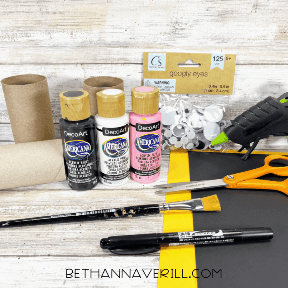 Craft supplies for penguin paper roll craft including paints, paper, scissors, glue gun, and googly eyes.