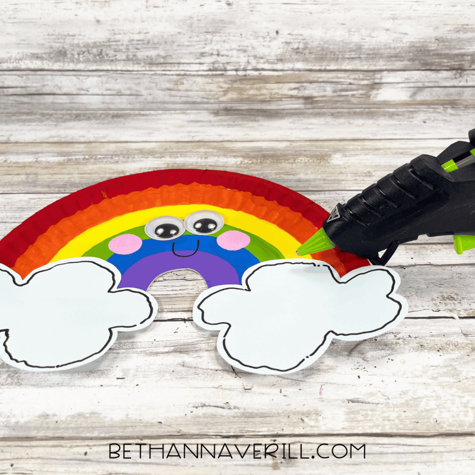 Rainbow paper plate with googly eyes, pink cheeks, and clouds being glued to each end.