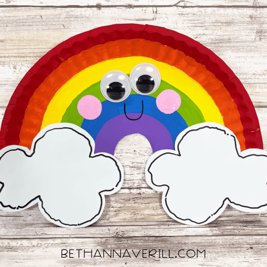 Finished rainbow paper plate craft with smiling face and white paper clouds on each side.
