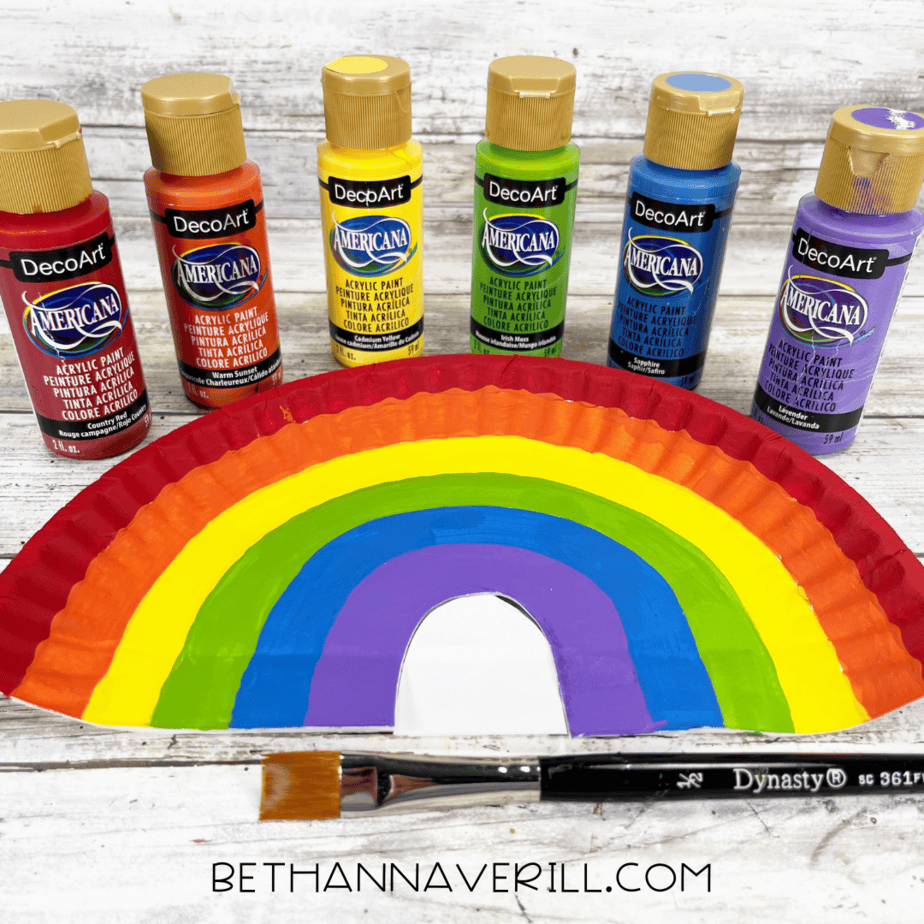 Half paper plate painted as a rainbow with bottles of paint behind it.