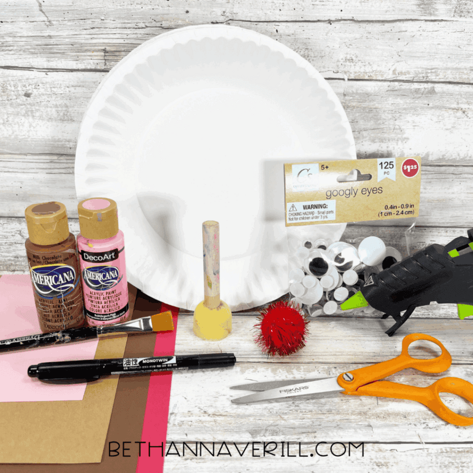 Rudolph reindeer craft supplies including paper plate, paints, construction paper, googly eyes, red pom pom, glue gun, and scissors.