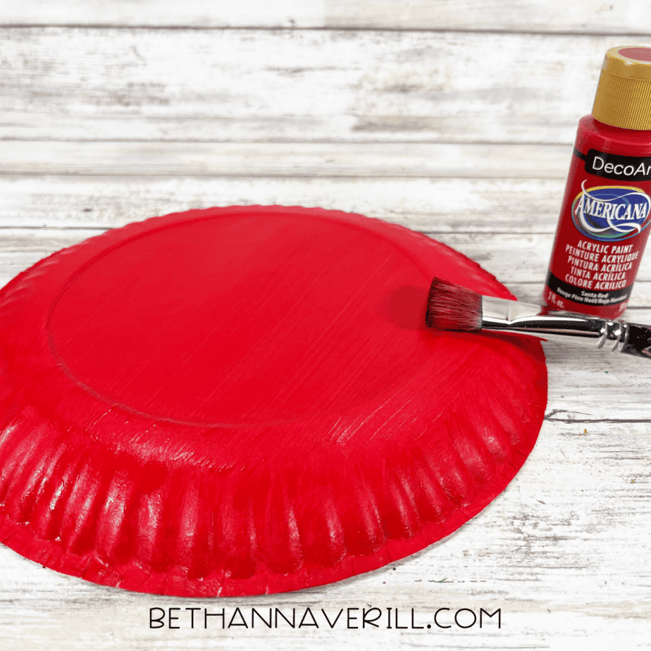 Paper plate painted red with paintbrush and red paint bottle beside it.