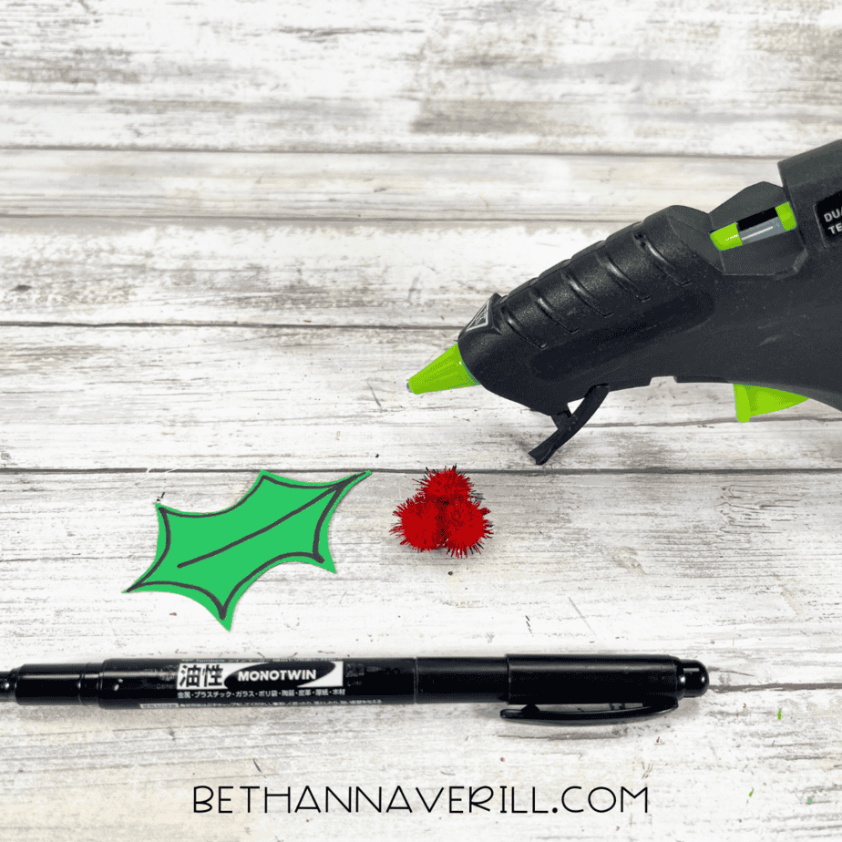 Cut-out holly leaves with black marker details and red pom-poms beside glue gun.