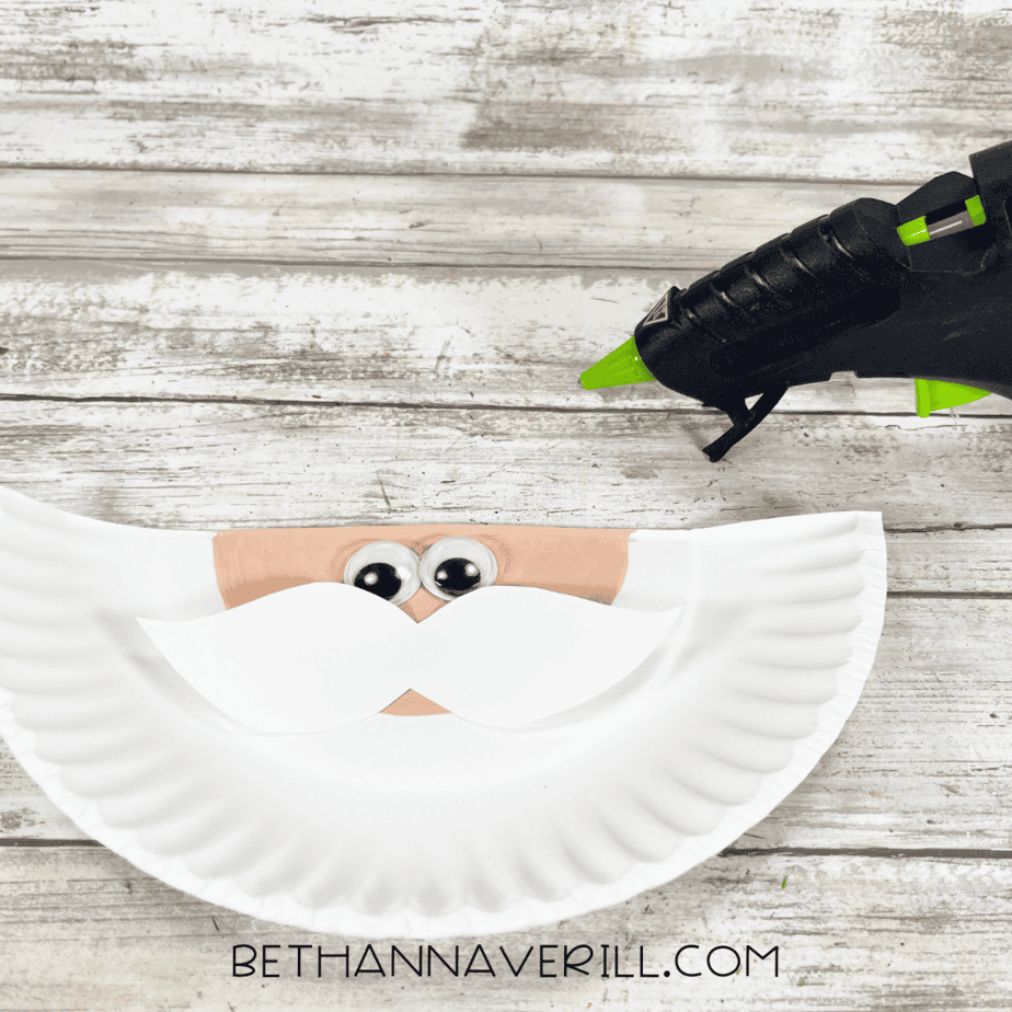 Half paper plate with peach face painted, googly eyes, and white mustache glued in place.