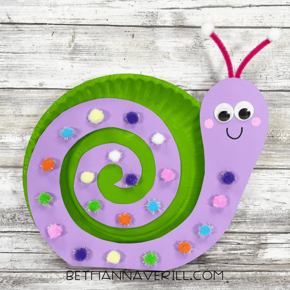 Finished paper plate snail craft with a purple spiral shell, pom poms, and smiling face.