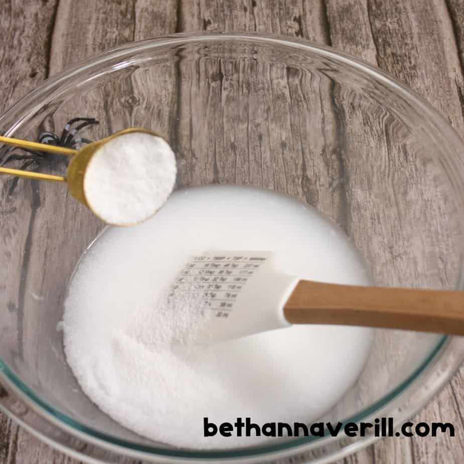 Another view of baking soda being poured into a glue and water mixture in a glass bowl, with a spatula resting inside. The spider toy theme continues in the background.