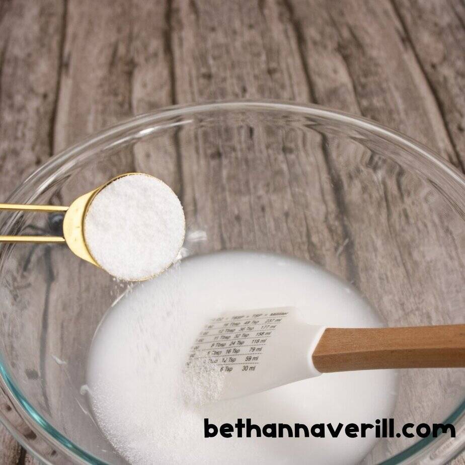 A spoonful of baking soda, is being added to a bowl of glue mixture with a spatula submerged in it. The surface has a wood grain background with a small toy spider in the corner.