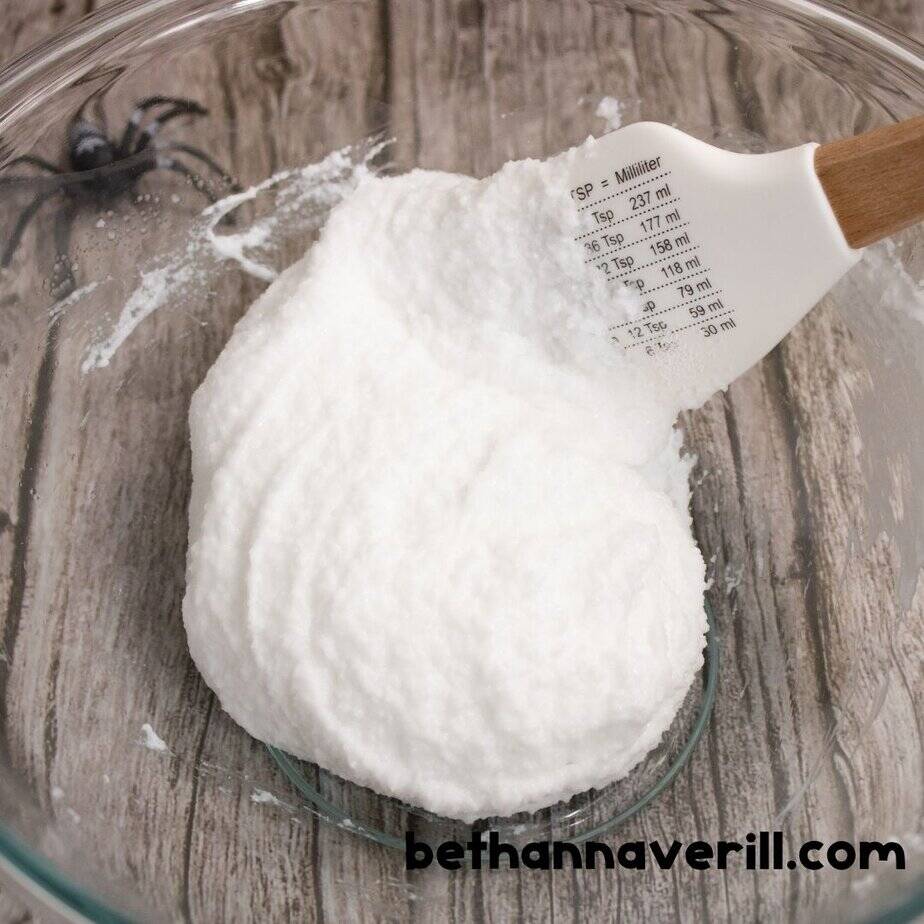 A close-up of thick white slime being stirred in a glass bowl with a spatula that has measuring conversions printed on it. A small plastic spider is visible in the background against the wood-textured surface.