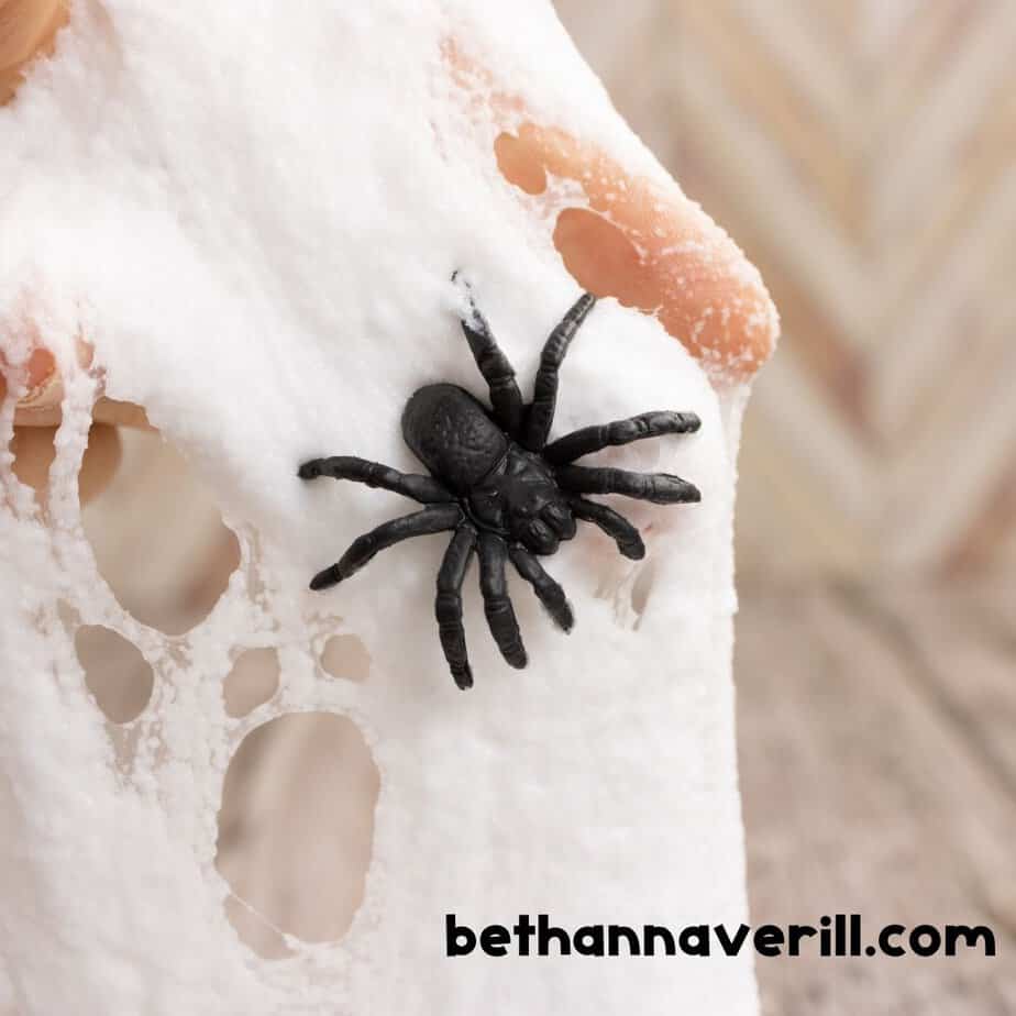 A stretched piece of fluffy white slime with a black toy spider embedded in it, held up by a hand. The slime has a web-like texture with holes.