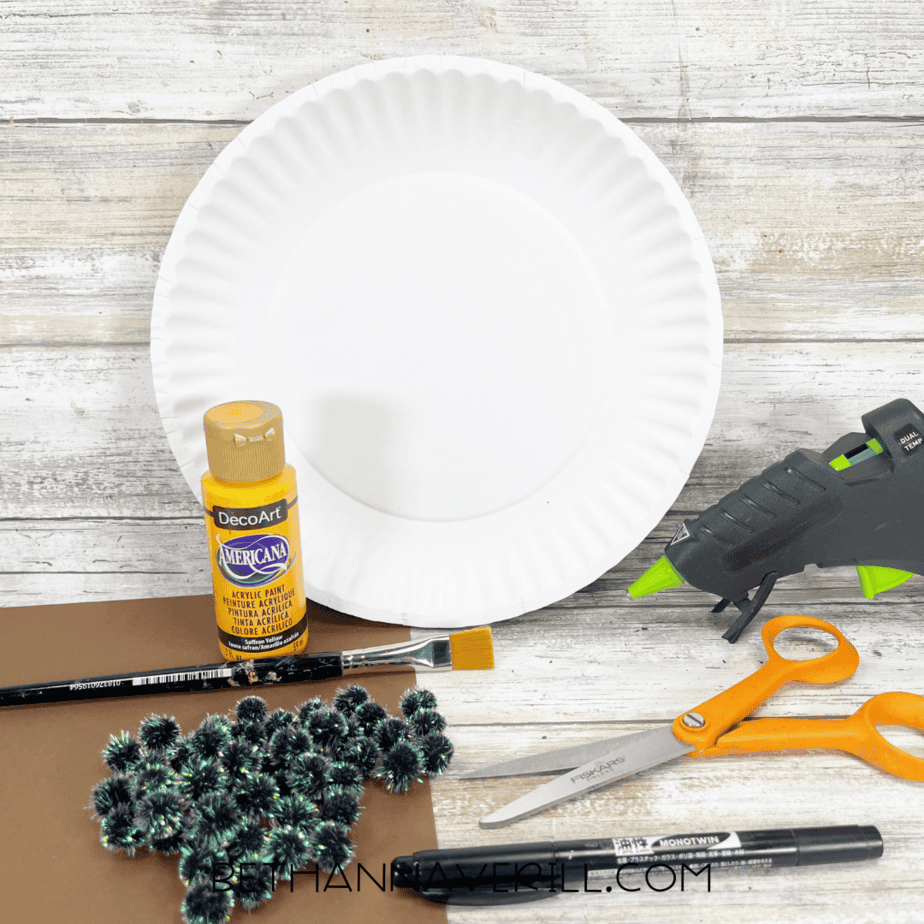 Sunflower paper plate craft supplies including paint, plate, pom-poms, glue gun, scissors, and construction paper.
