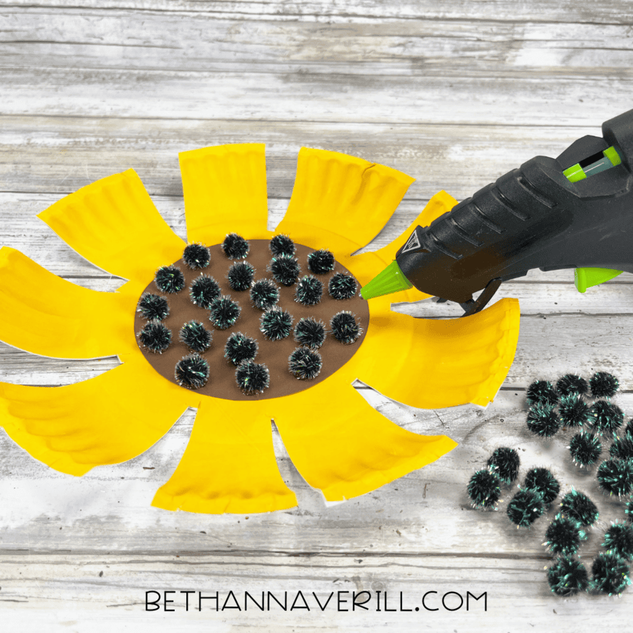 Glue gun attaching black pom-poms to the brown center of the sunflower.