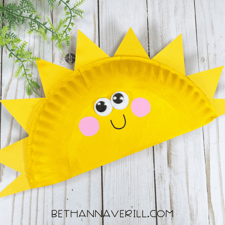 Completed sunshine paper plate craft with googly eyes, pink cheeks, and sun rays, displayed on a wooden surface with greenery.
