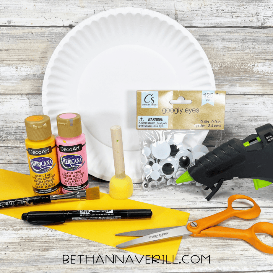 Craft supplies including a paper plate, yellow and pink paint, googly eyes, scissors, glue gun, and yellow paper.