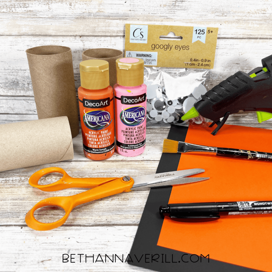 Materials for tiger paper roll craft including paint, cardboard rolls, googly eyes, scissors, glue gun, and construction paper.