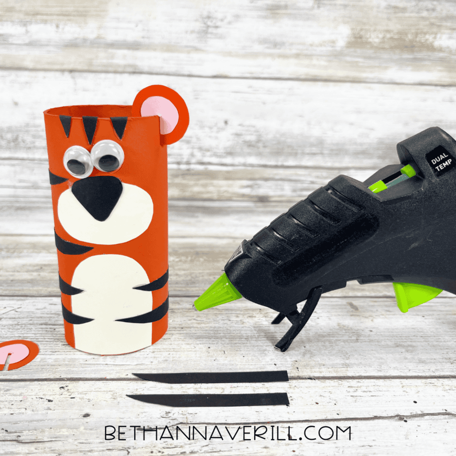 Tiger paper roll partially assembled with googly eyes, nose, and stripes being glued on.